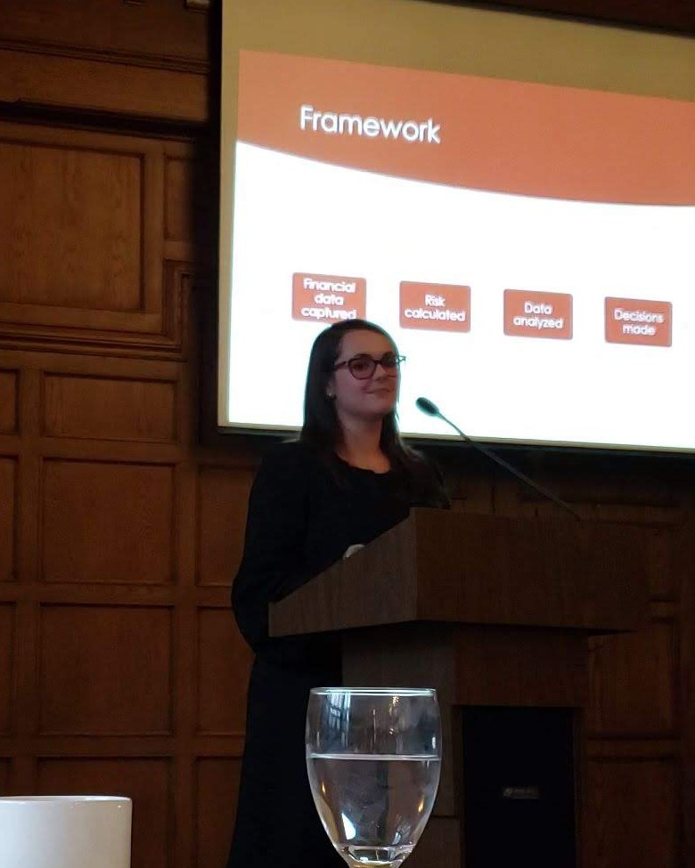 Galina Fendikevich speaking from a wooden lectern at a New York State Society of CPAs keynote, with a slide titled Framework showing financial data captured, risk calculated, data analyzed and decisions made projected behind her