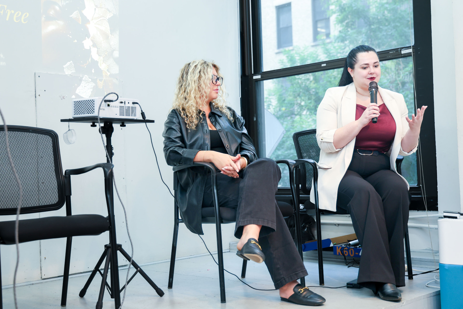 Two panelists seated on a low stage with a projector beside them, Galina Fendikevich holding a microphone and gesturing