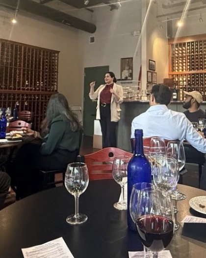 Galina Fendikevich standing and speaking to a seated audience in a wine tasting room, floor-to-ceiling wine racks behind her