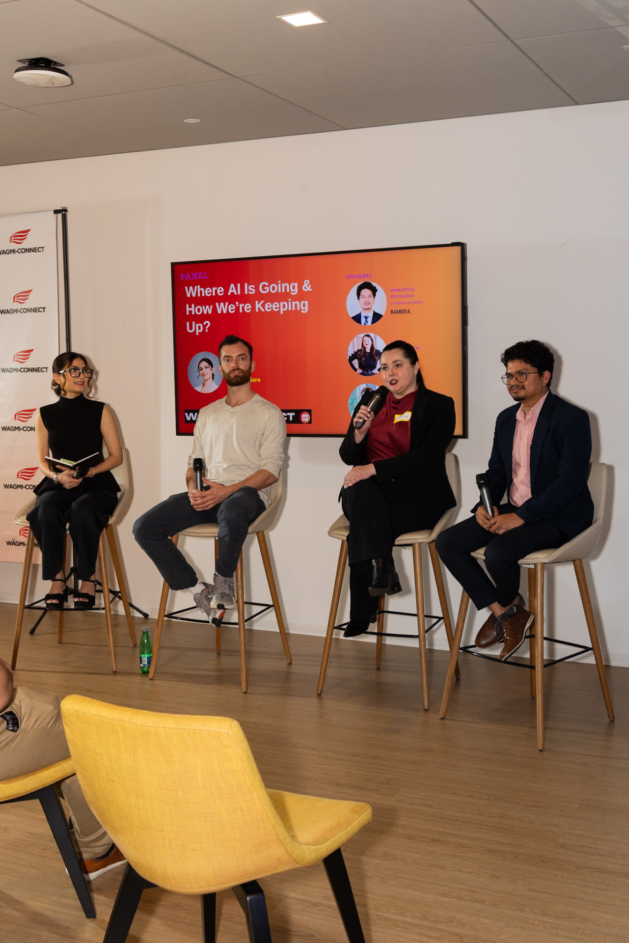 Four panelists seated on tall stools against a white wall with the panel topic displayed on a screen behind them