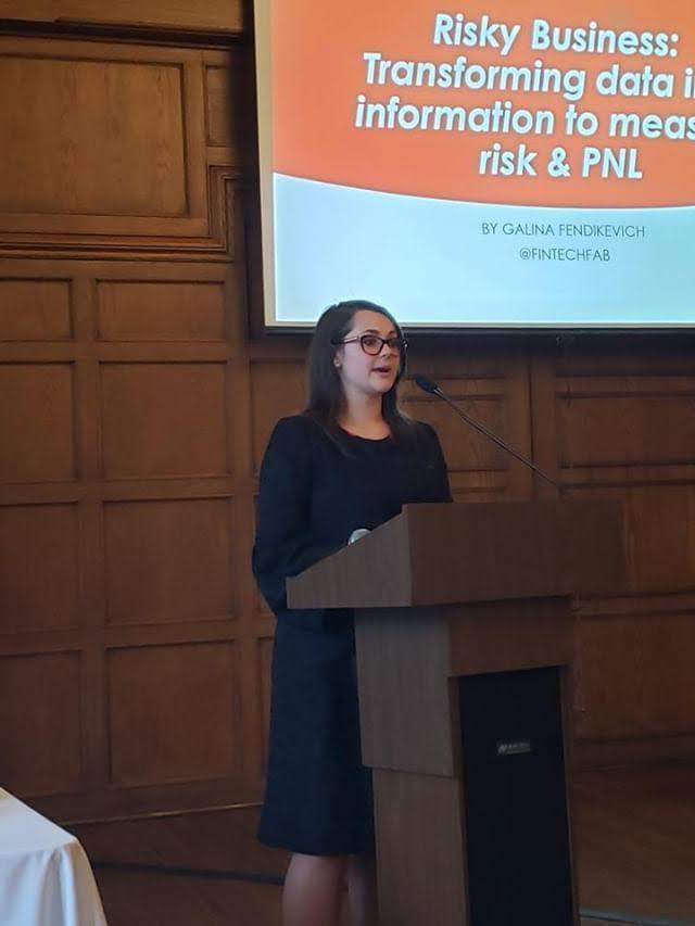 Galina Fendikevich speaking into a microphone at a lectern during her New York State Society of CPAs keynote, with the slide Risky Business: transforming data into information to measure risk and PNL behind her